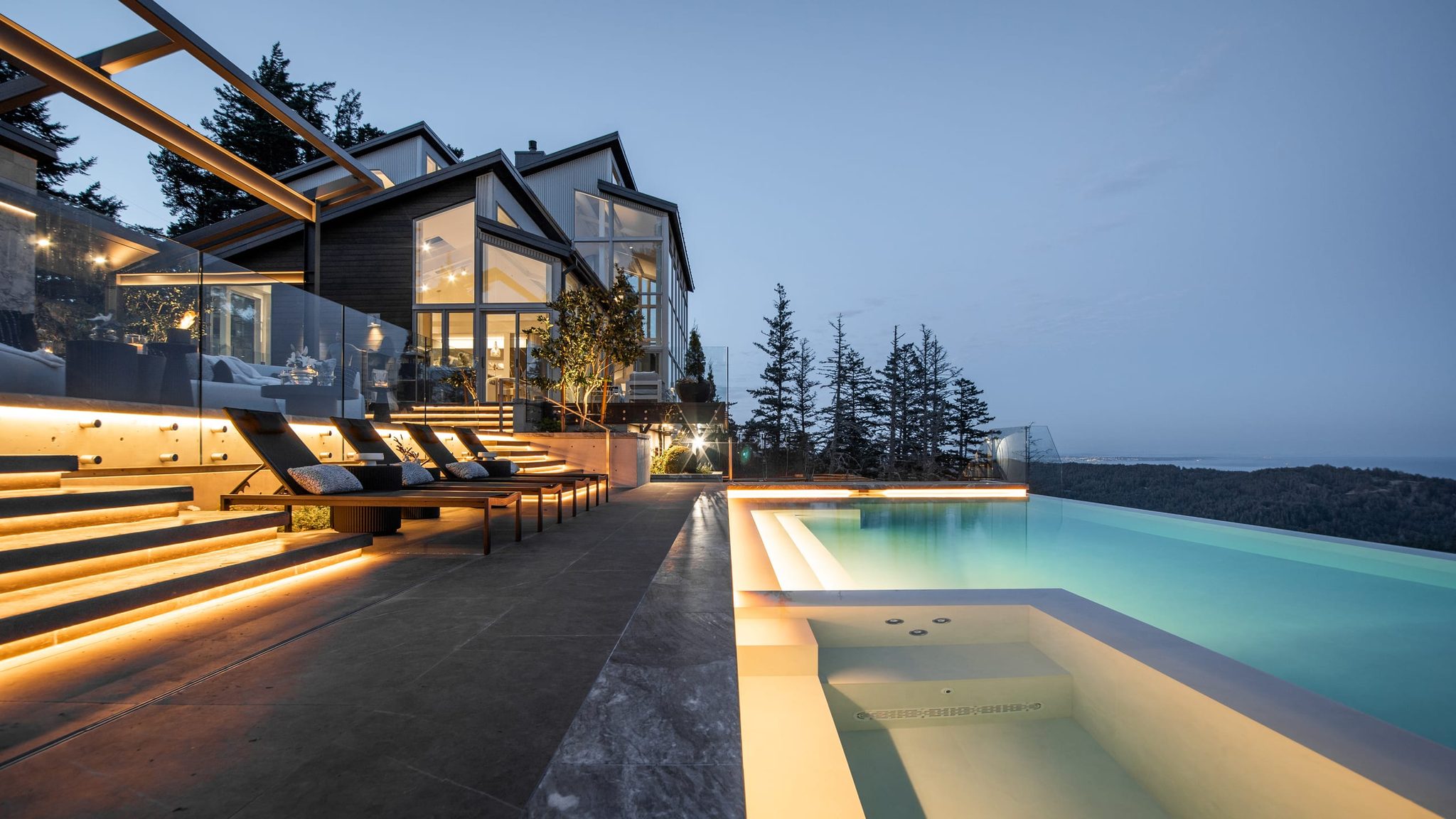 Modern custom home and its pool lit in the evening, with forested hills beyond