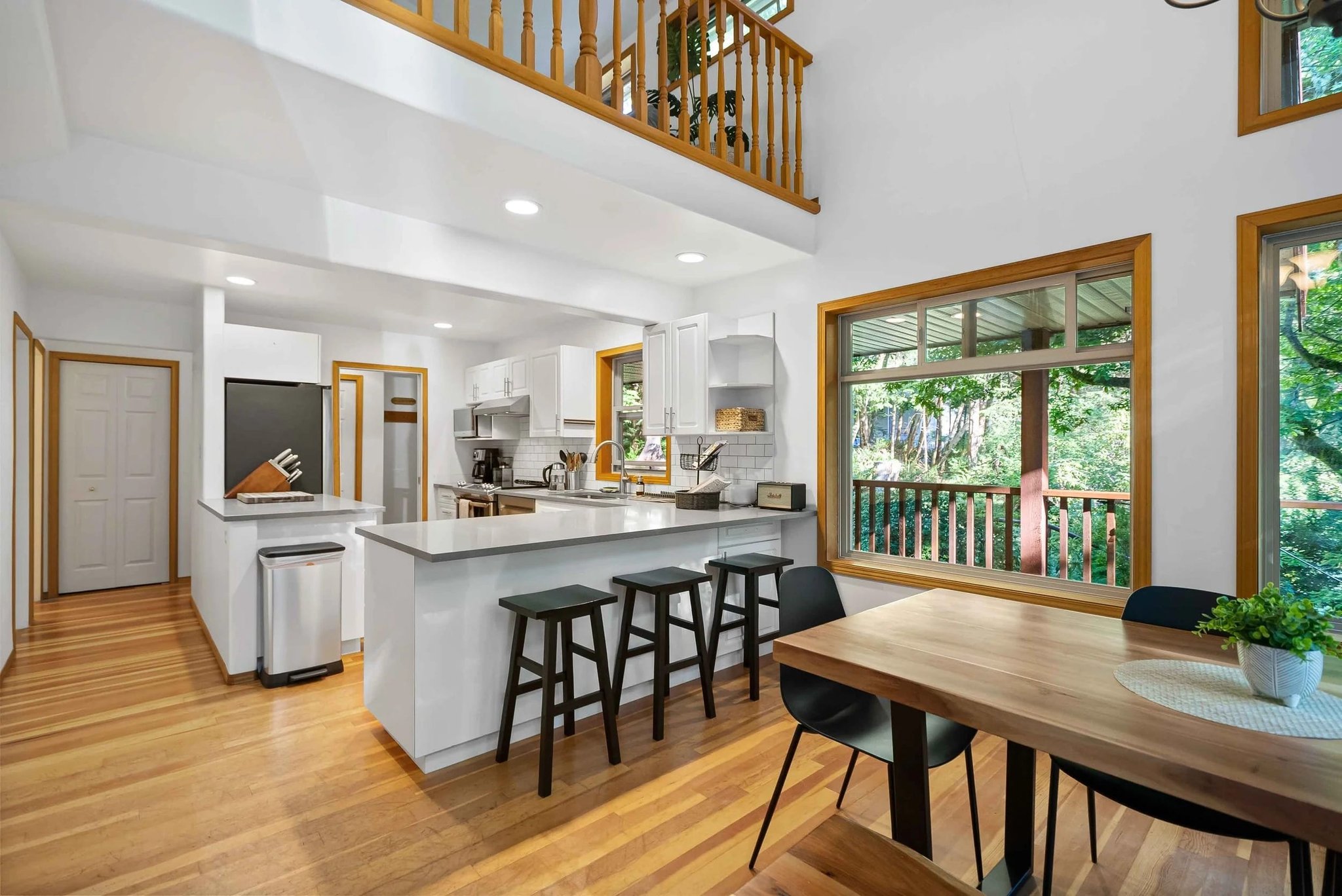 Vaulted open-concept kitchen and dining area with a loft and wood-trimmed windows overlooking trees